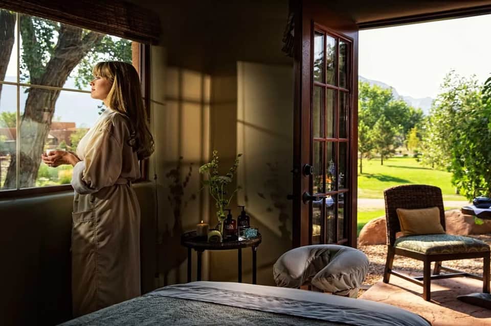 Woman in spa robe looking out at lush green grounds through open glass doors from bedroom