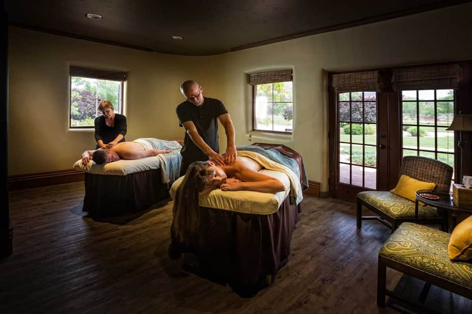 Spa massage room with two therapists treating clients on massage tables with windows overlooking grounds