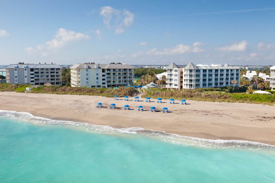 Beachfront resort with multiple pastel-colored buildings and blue umbrellas on sandy beach
