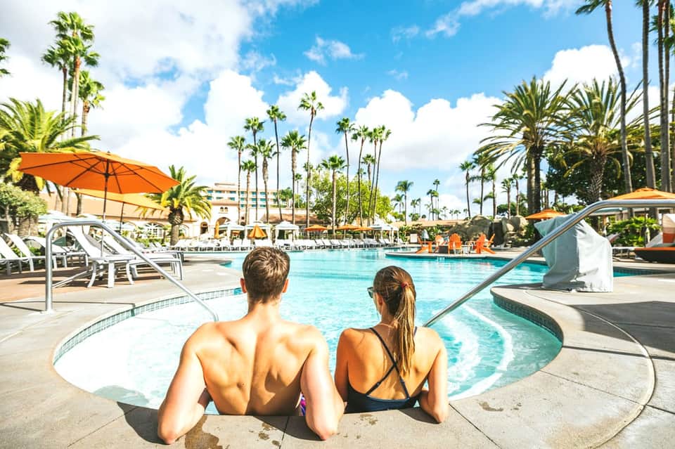 Two guests sitting at edge of resort pool with palm trees, lounge chairs, and orange umbrellas