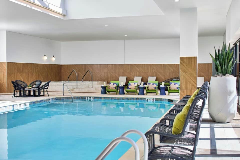 Modern indoor pool with high ceilings, wooden wall accents, green lounge chairs, black wicker seating, and skylights