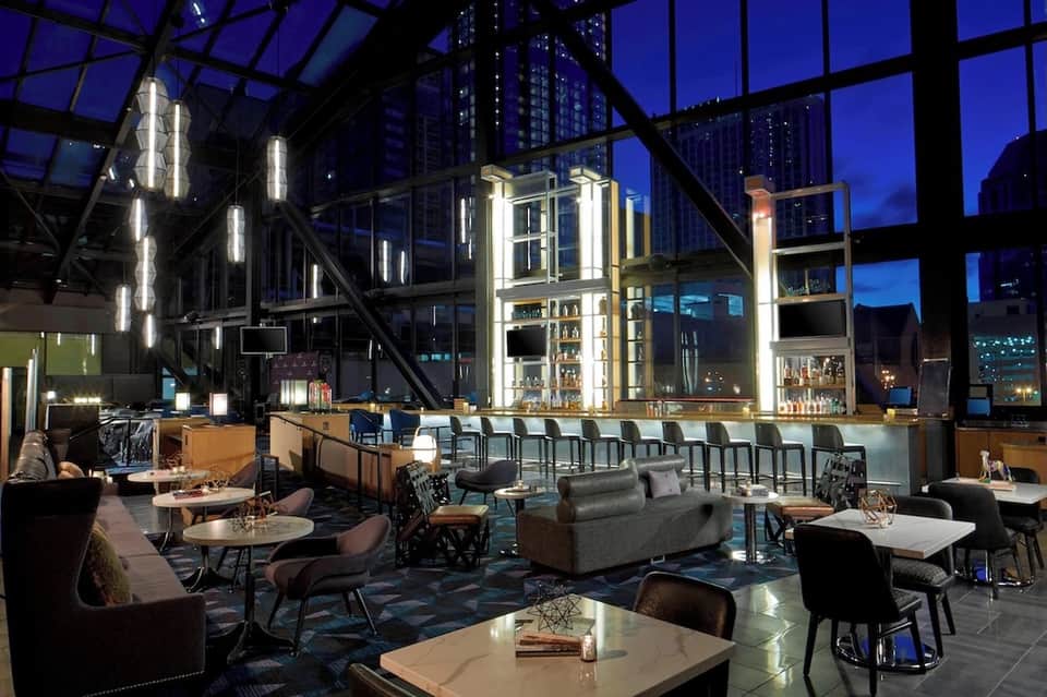 Contemporary rooftop bar with glass ceiling, city skyline views, bar seating, and dining tables