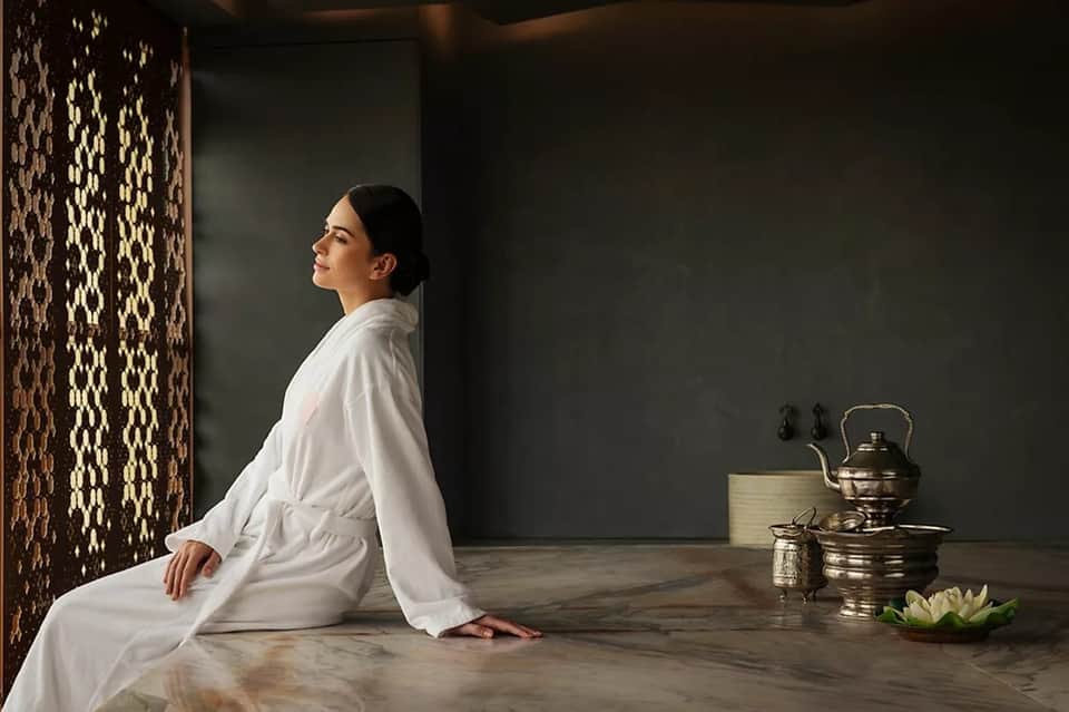 Woman in white robe sitting on marble spa floor with decorative screen and traditional tea service
