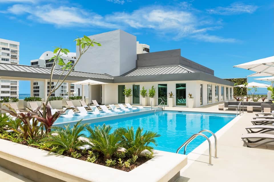 Resort pool and terrace with white umbrellas, tropical landscaping, and ocean view under blue sky