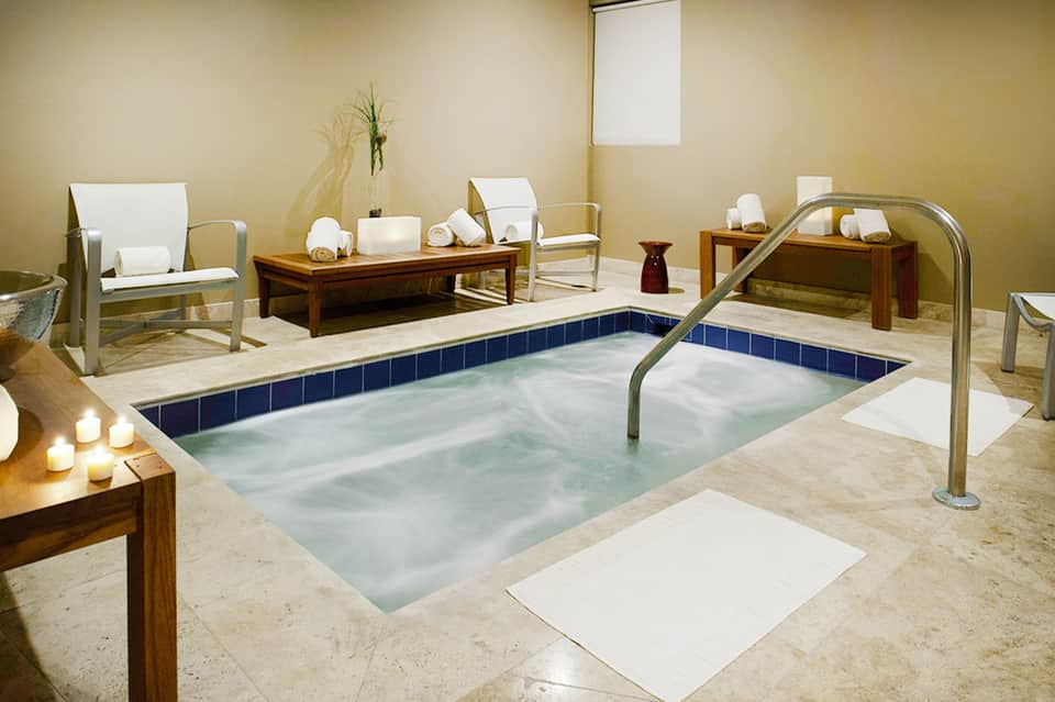 Spa indoor pool with blue tile accents, white lounge chairs, and wooden tables with rolled towels