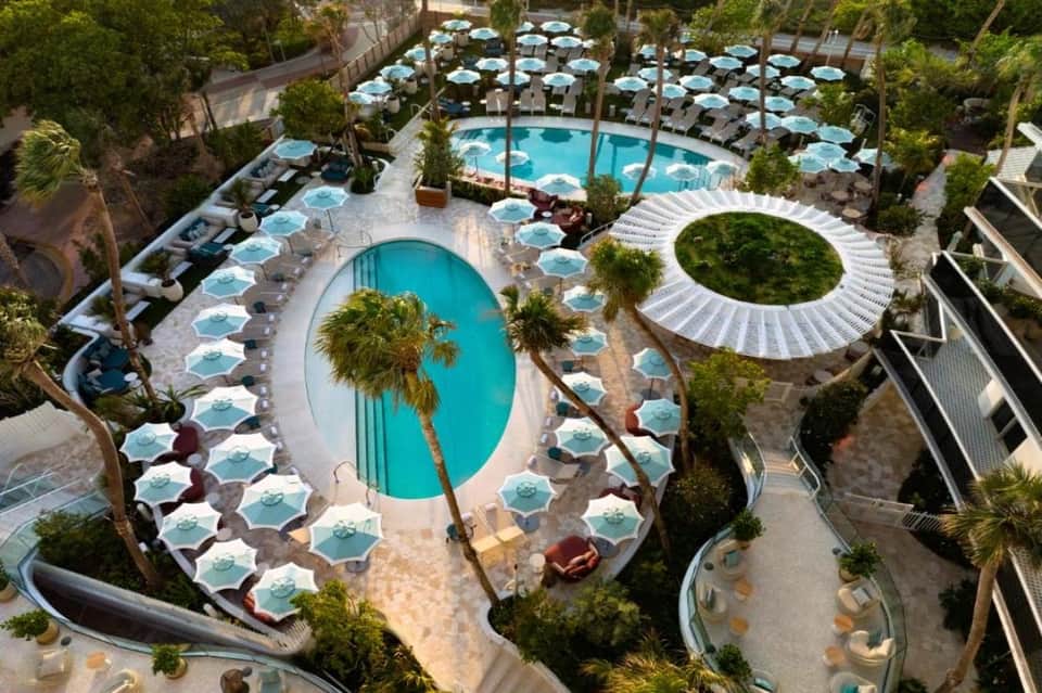 Aerial view of resort pool complex with turquoise water, blue umbrellas, loungers, and palm trees
