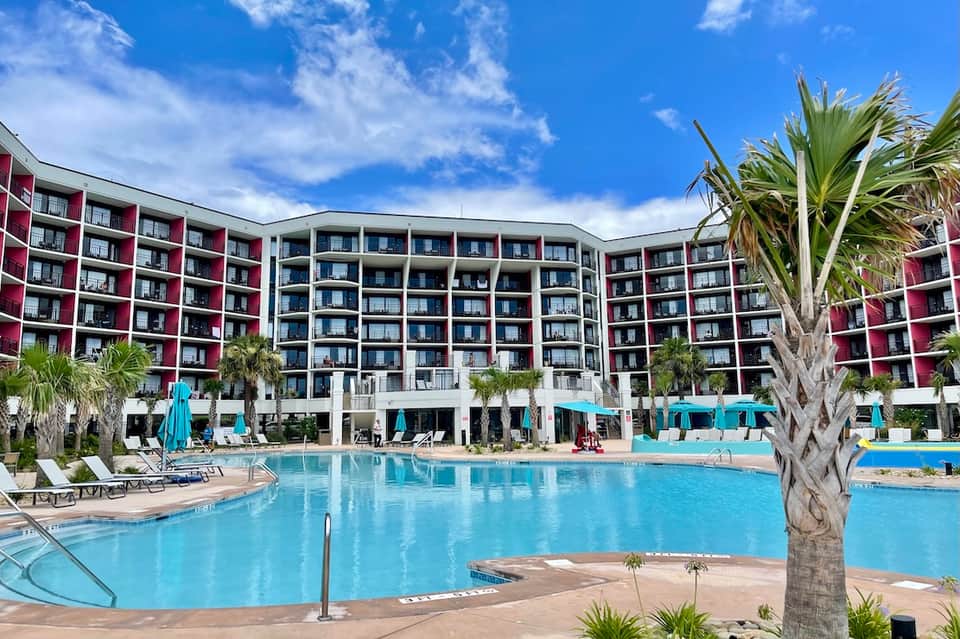 Large resort pool with blue water, modern multi-story hotel building, lounge chairs, and turquoise umbrellas