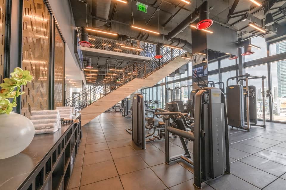 Modern gym with equipment and city views through large windows, mezzanine level visible