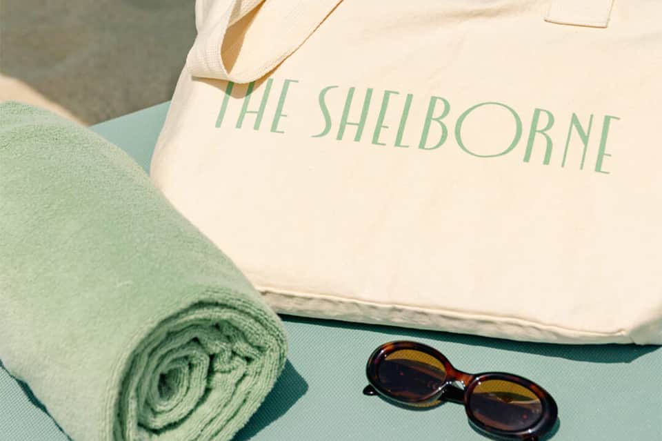 The Shelborne branded canvas tote bag with rolled green towel and sunglasses on turquoise surface