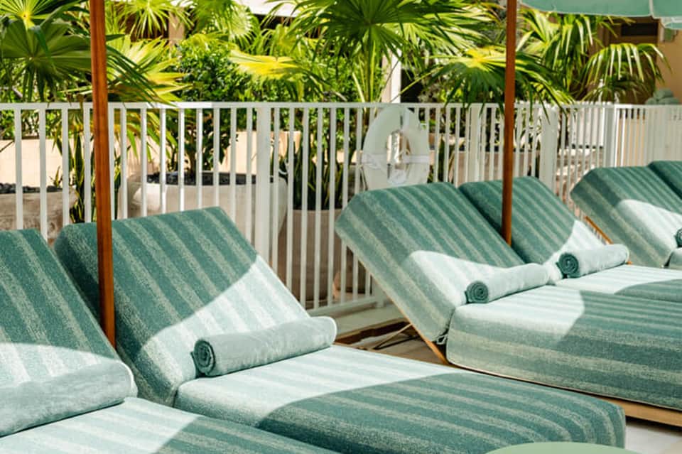 Striped green and white lounge chairs with rolled towels under pergola with tropical palm plants