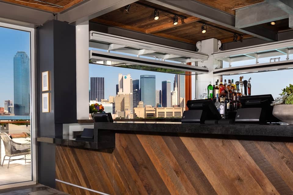 Modern rooftop bar with dark wood counter, floor-to-ceiling windows showcasing urban skyline, bottle display, and wooden ceiling