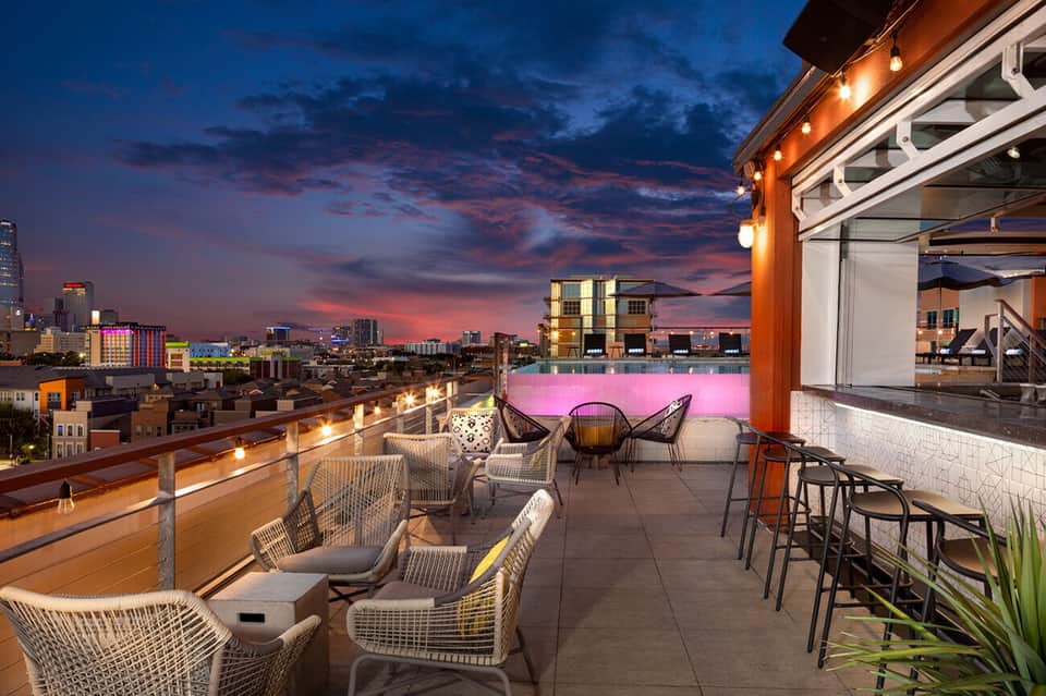Rooftop bar and lounge at sunset with pink pool, woven chairs, and dramatic purple and orange sky