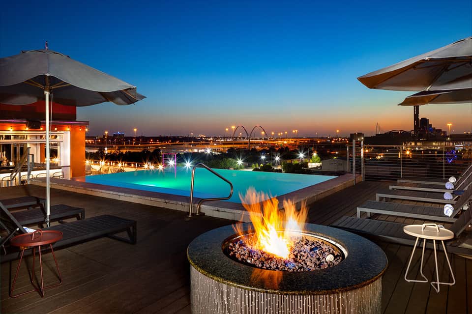 Rooftop pool with glowing blue water, fire pit, umbrellas, and city skyline at dusk