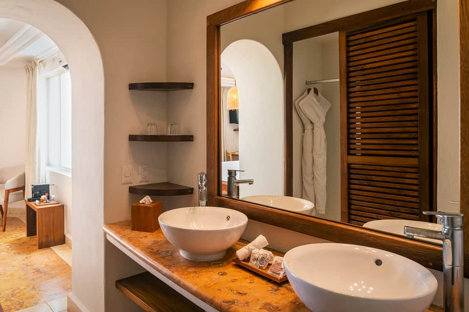 Luxury bathroom with dual white vessel sinks on cork countertop, wooden frame mirror, arched alcove, and louvered cabinet