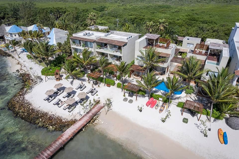 Aerial view of beachfront resort with white buildings, palm trees, sandy beach, and turquoise ocean