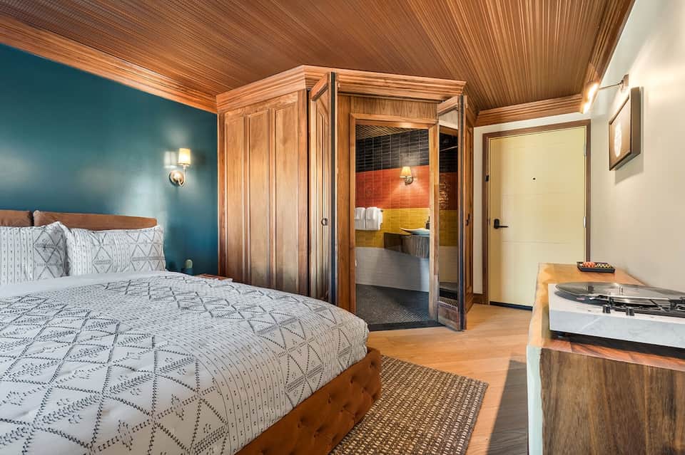 Modern bedroom with teal accent wall, wooden furnishings, and colorful tiled bathroom visible