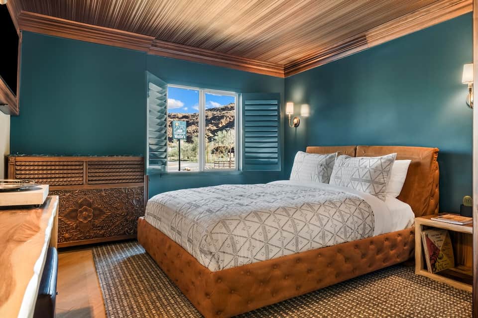 Modern hotel bedroom with teal walls, wooden ceiling, leather bed, and mountain view