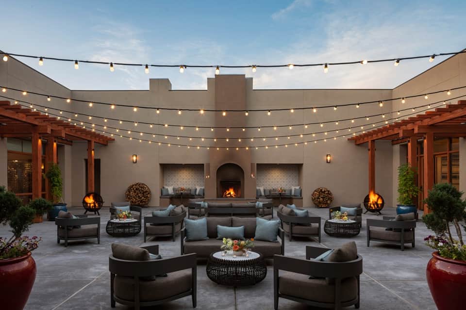 Outdoor courtyard lounge with fireplace, string lights, dark furniture, and contemporary fire pits
