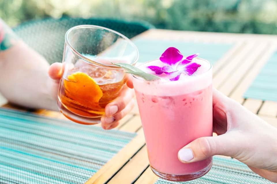 Two colorful tropical cocktails by a poolside—pink drink with orchid garnish and amber drink with citrus slice