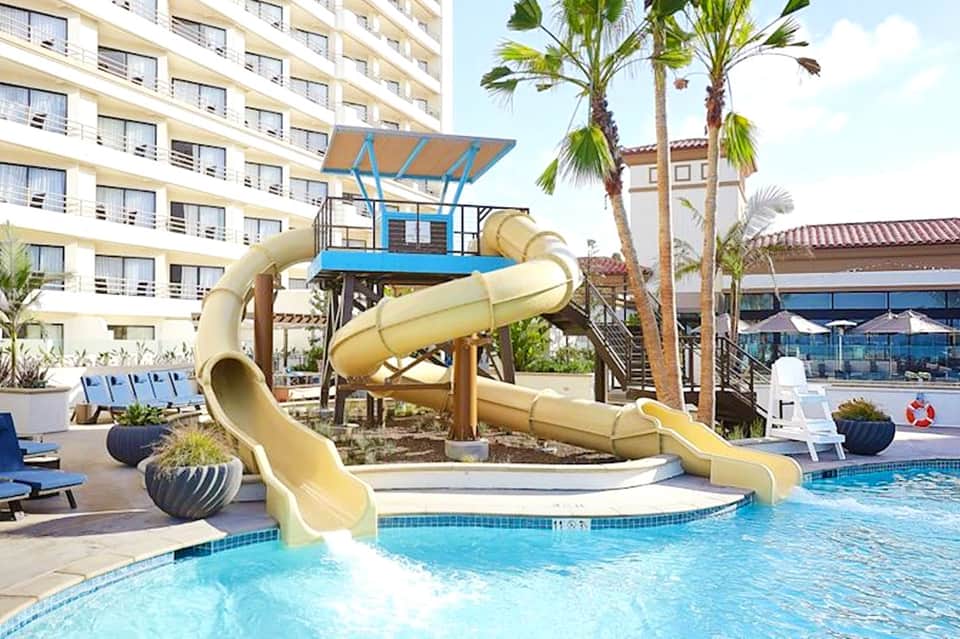 Resort pool with yellow waterslide, palm trees, and multi-story hotel building in background
