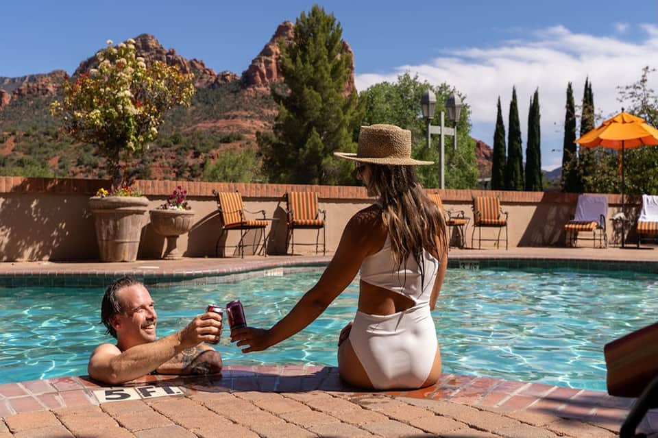 Resort pool with mountain backdrop, woman at poolside, desert scenery, orange umbrella, and striped lounge chairs