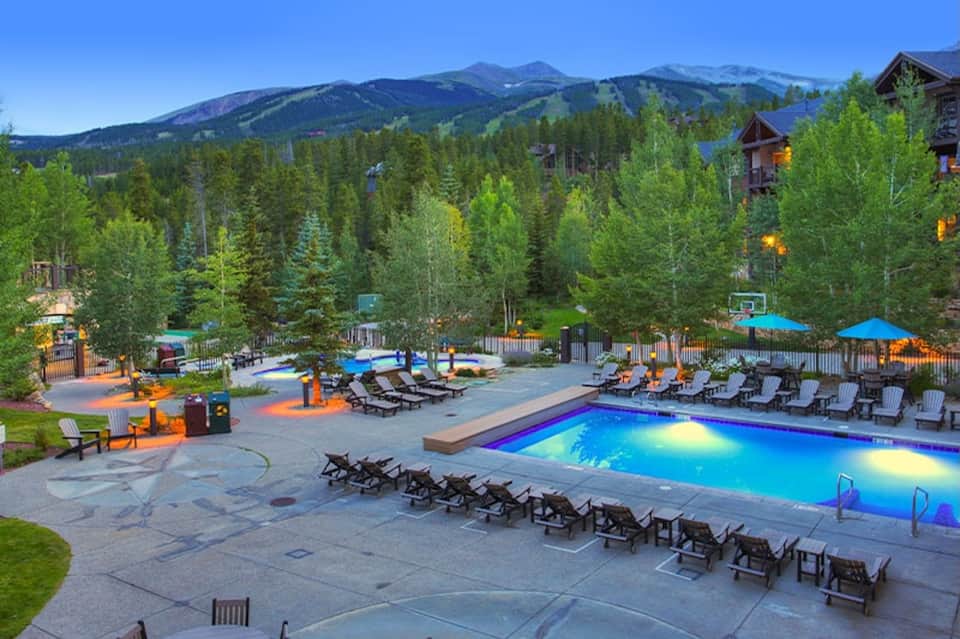 Aerial view of illuminated resort pool at dusk with lounge chairs and mountainous forest backdrop