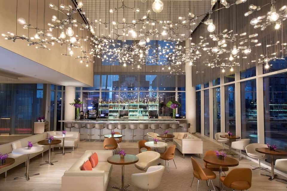 Modern hotel lounge with illuminated bubble ceiling installation, bar area, and seating overlooking city at dusk