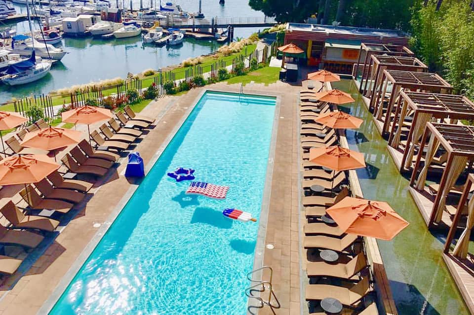 Rectangular swimming pool lined with lounge chairs and orange umbrellas, overlooking a marina with sailboats.
