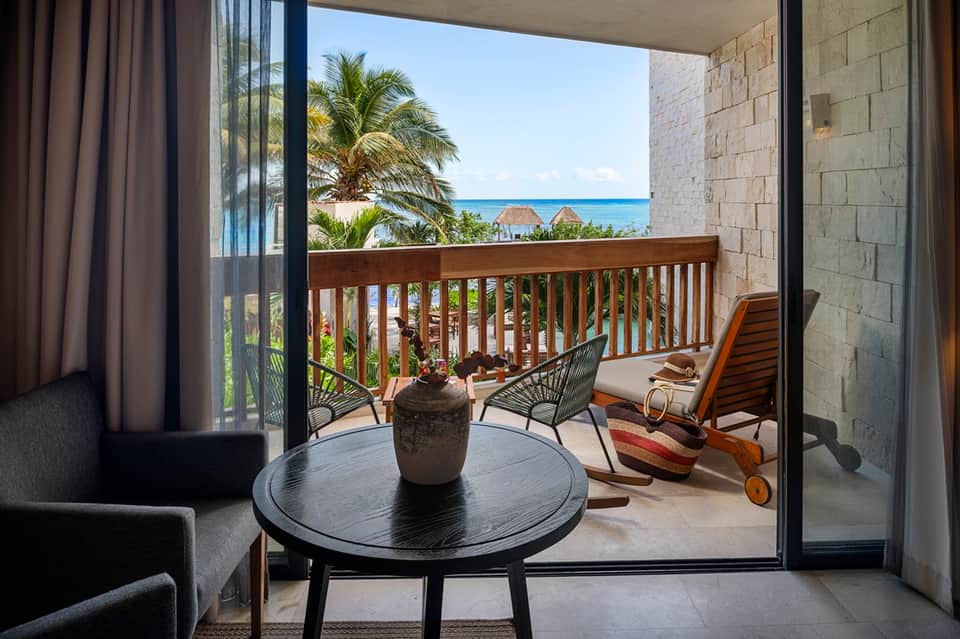 Beachfront balcony with ocean view, palm trees, rattan chairs, and sliding glass doors from bedroom