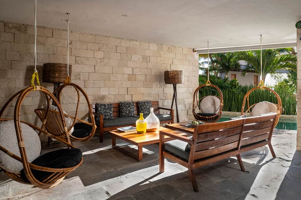 Outdoor covered lounge with hanging woven chairs, wooden furniture, and stone accent wall overlooking tropical garden.