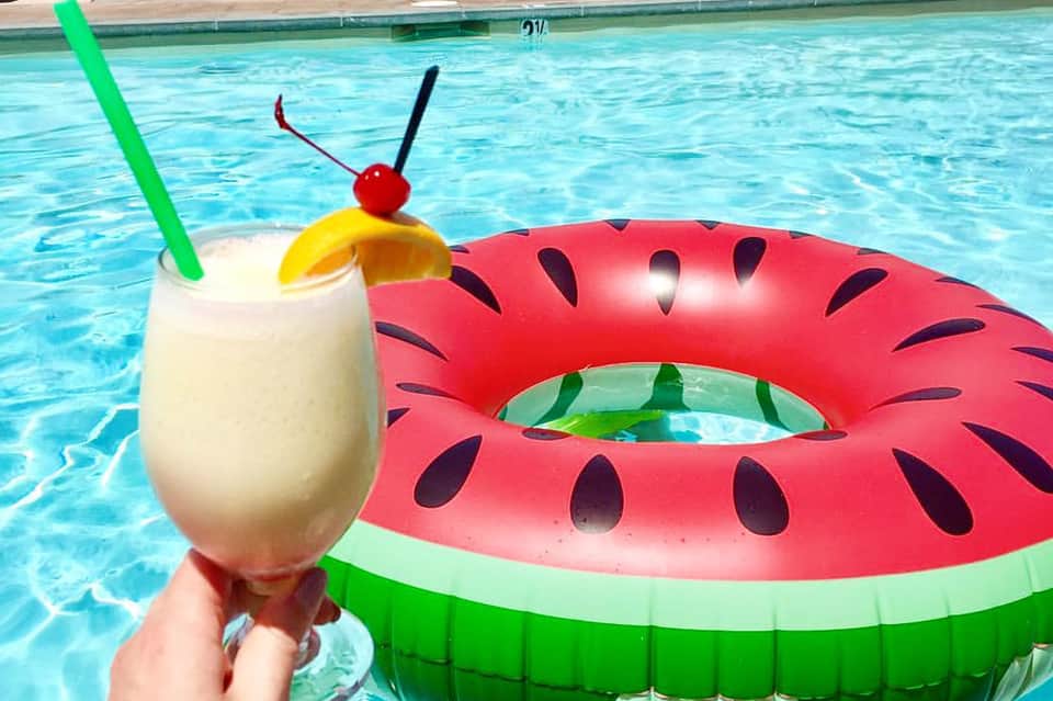 Tropical cocktail with green straw and cherry garnish beside watermelon pool float in swimming pool
