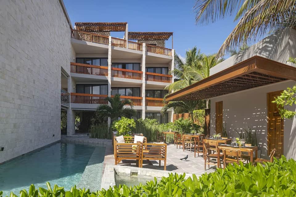 Modern resort courtyard with pool, wooden lounge chairs, pergola shade structure, and palm trees