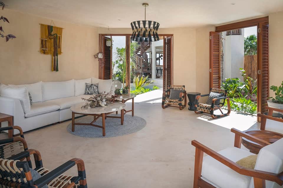 Bright open-concept living room with cream sofa, wooden furnishings, large windows opening to tropical courtyard.