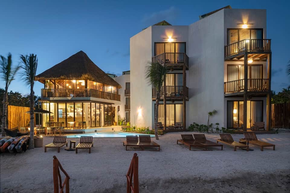 Beachfront resort at dusk with illuminated buildings, pool, loungers, and thatched-roof structures