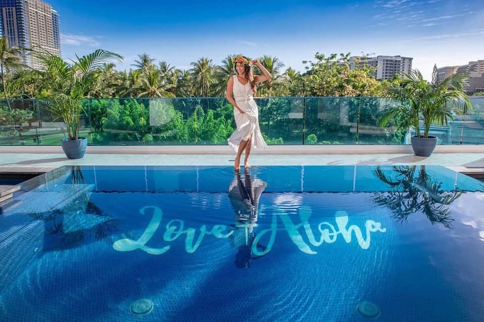 Woman standing on pool deck beside blue water with 'Aloha' text