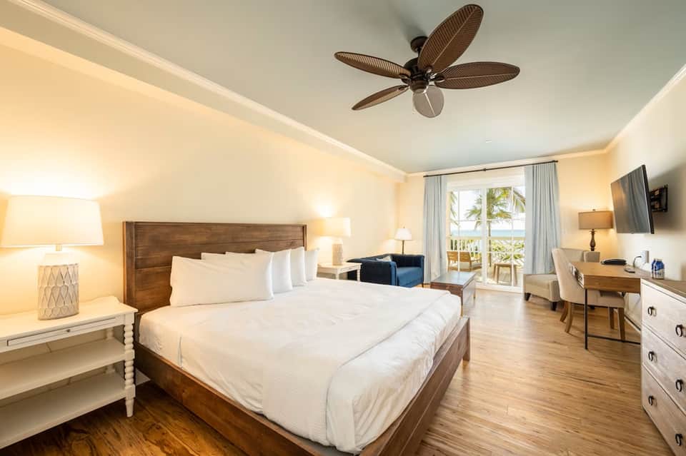 Bright hotel bedroom with king bed, wood furniture, ocean view balcony, and ceiling fan