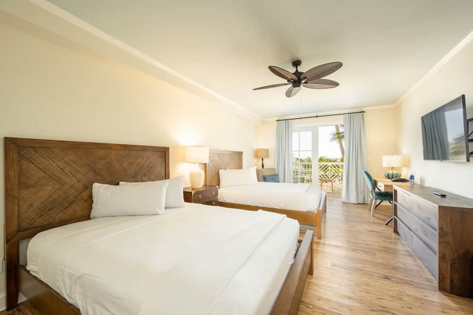 Tropical resort bedroom with two queen beds, wood headboards, balcony access, and ocean-view windows