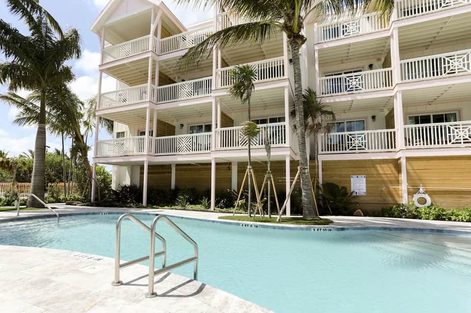 Three-story residential building with white railings and palm trees, featuring screened ground level and pool access