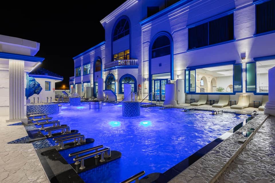 Illuminated resort pool at night with blue lighting, loungers, and modern architectural building facade