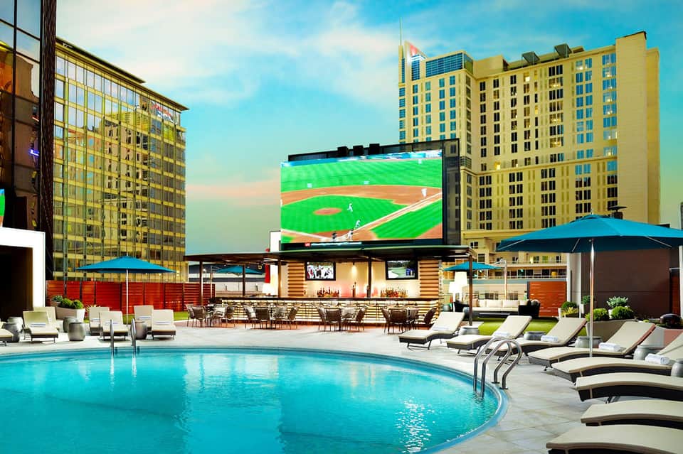 Resort rooftop pool with lounge chairs, bar area, and large sports stadium display, surrounded by yellow high-rise buildings