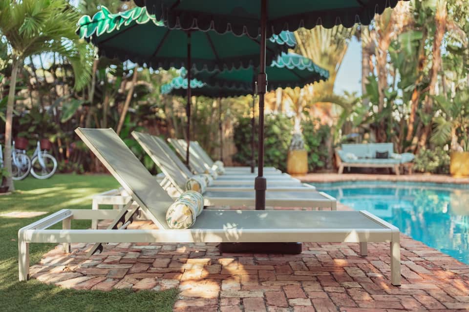 Lounge chairs under green umbrellas beside crystal-blue pool with palm trees and resort buildings