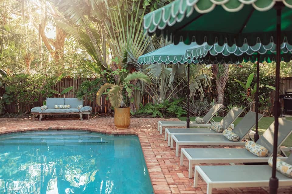 Residential pool with brick patio, lounge chairs under green awning, palm trees, and garden seating