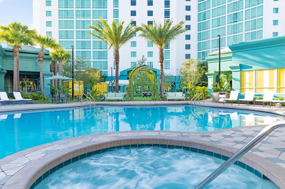 Resort pool area with turquoise water, hot tub, palm trees, lounge chairs, and modern hotel building