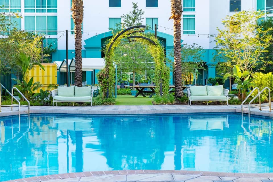 Resort pool with blue water, white lounge chairs, green archway trellis, and turquoise building facade