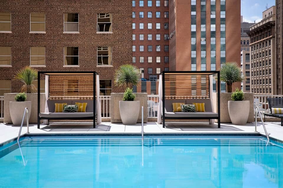 Urban rooftop pool with bright blue water, black cabanas with yellow pillows, and downtown city buildings