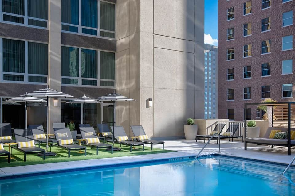 Urban rooftop pool surrounded by striped umbrellas, lounge chairs, and modern city buildings
