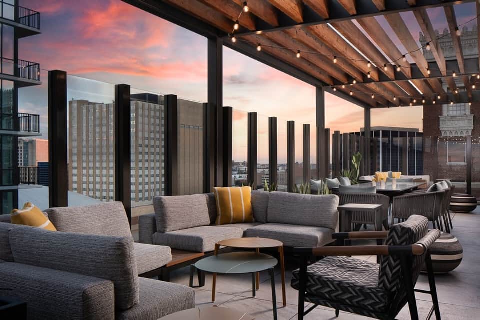 Modern rooftop lounge at sunset with gray seating, wood pergola, city skyline views, and accent lighting