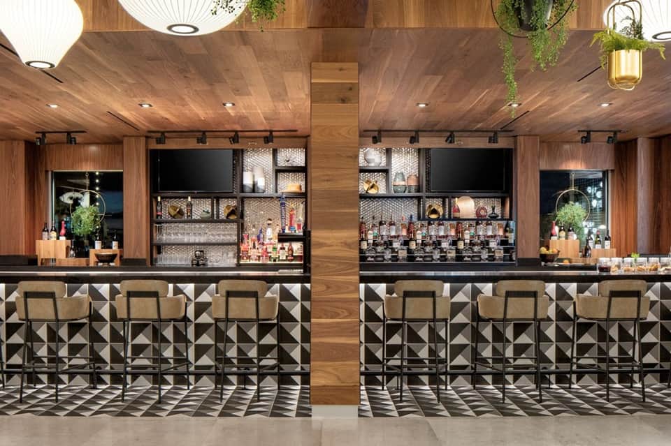 Modern hotel bar with wooden ceiling, geometric patterned counter, and tiered shelving displays