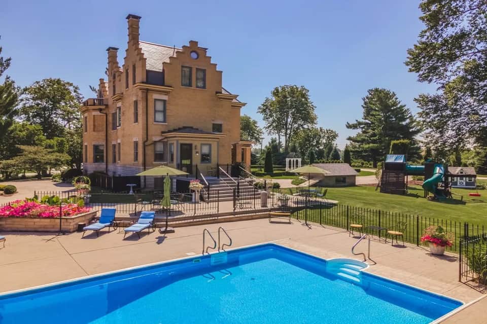 Historic brick mansion with pool in foreground, manicured lawn, gardens, and playground equipment