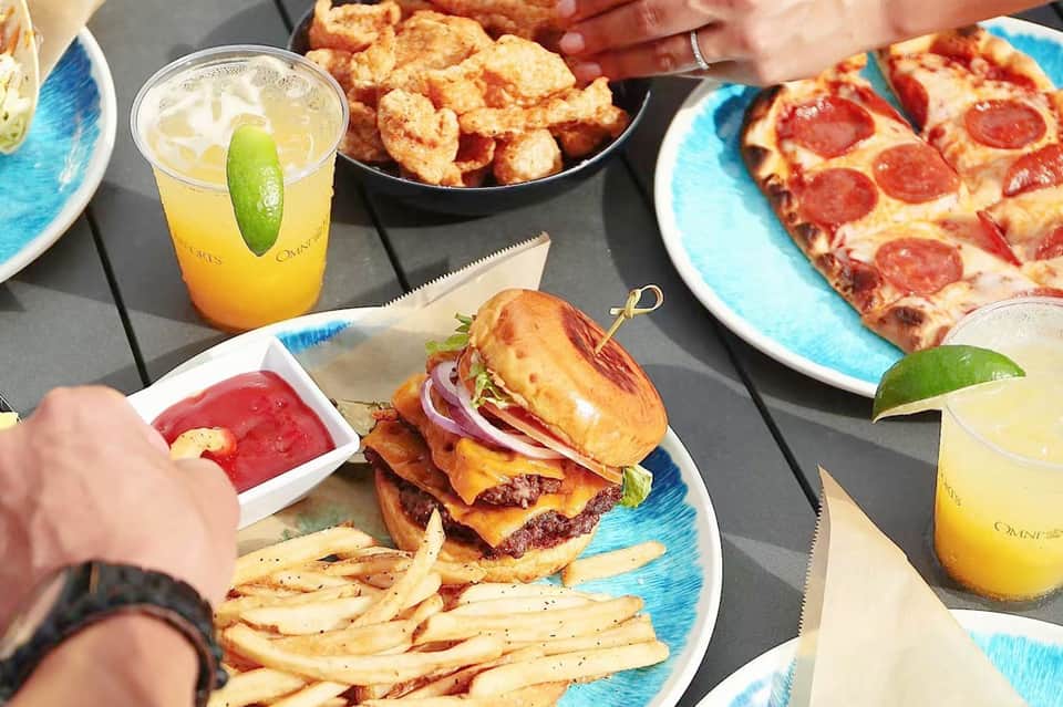 Casual dining spread with burger, fries, fried shrimp, pizza, and citrus cocktails at hotel restaurant